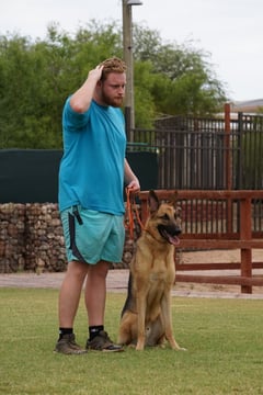 Partners Dogs trainer in a teal shirt standing beside a German Shepherd holding a calm sit-stay during an outdoor obedience training session