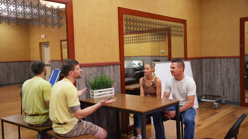 Two Partners Dogs trainers in yellow shirts explain program options to a couple seated at a table during an enrollment consultation.