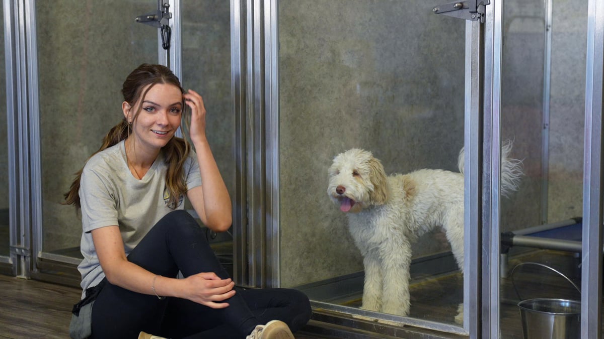 A smiling Partners Dogs staff member sits beside a happy Goldendoodle in a clean indoor boarding suite